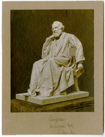 Photograph of a statue of Henry Wadsworth Longfellow on a pedestal. Longfellow wears a suit and a professor/graduation gown. He has chin-length hair and a full beard. He leans his chin on his right hand and holds a book casually in his left, his index finger keeping his page for him.