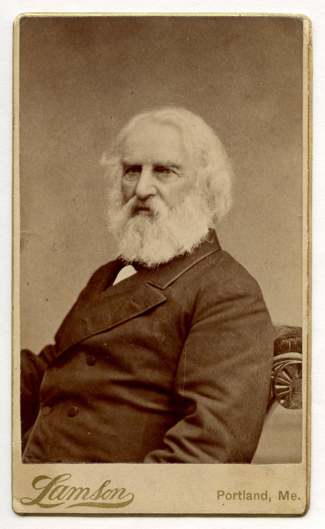 Henry Wadsworth Longfellow is seated three-quarter view to his right. He wears a double-breasted jacket with two buttonholes visible in his left lapel. A carved rosette of the chair is visible right. Longfellow's chin-length hair is fully white, as is his full beard.