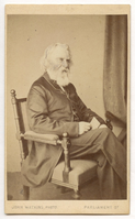 Almost full-length view. Henry Wadsworth Longfellow seated at three-quarter to viewer's right in Aesthetic-style armchair. Drapery is visible in the right background. He holds a piece of paper in his right hand. Longfellow wears a jacket and has white chin-length hair and a long white beard.