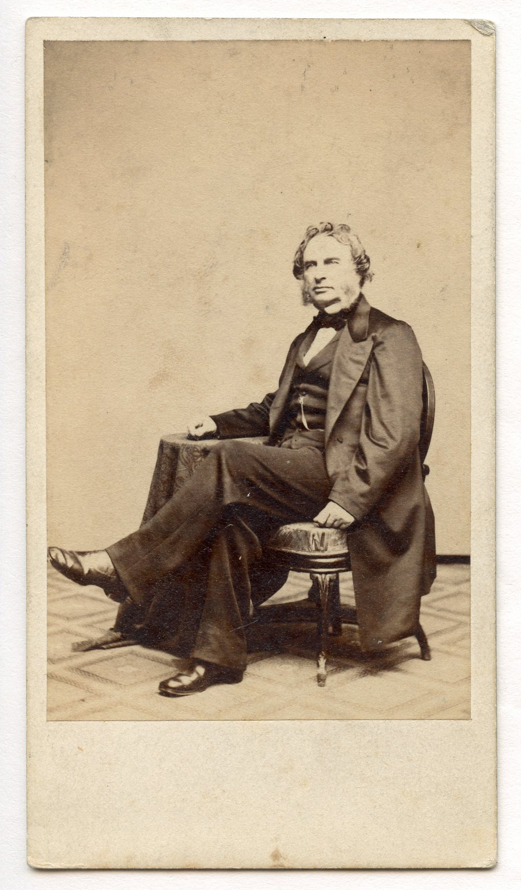 Henry Wadsworth Longfellow shown full-length, three-quarter view sitting with his right arm resting on table. The floor covering has a diamond-shaped pattern. A portion of the heavily decorated upholstery on the chair seat is visible. Longfellow has ear-length hair and graying sideburns.