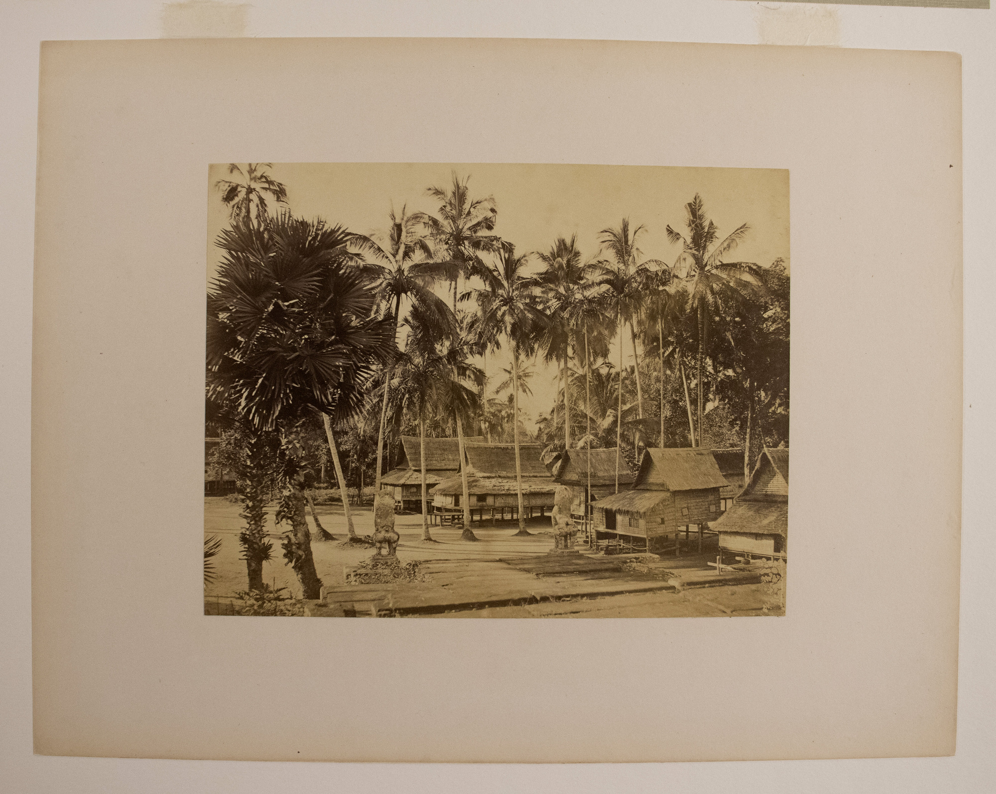 Sepia photograph with an inscription representing seven houses elevated on stilts, palm trees among them, and two statues flanking a road along the houses.