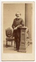 Henry Wadsworth Longfellow stands to the side of the balustrade with his left arm leaning on the top of its small pillar. An upholstered side chair is to the left and a Greek column is to the right in the background. Longfellow's hair is ear-length and he sports a graying beard.