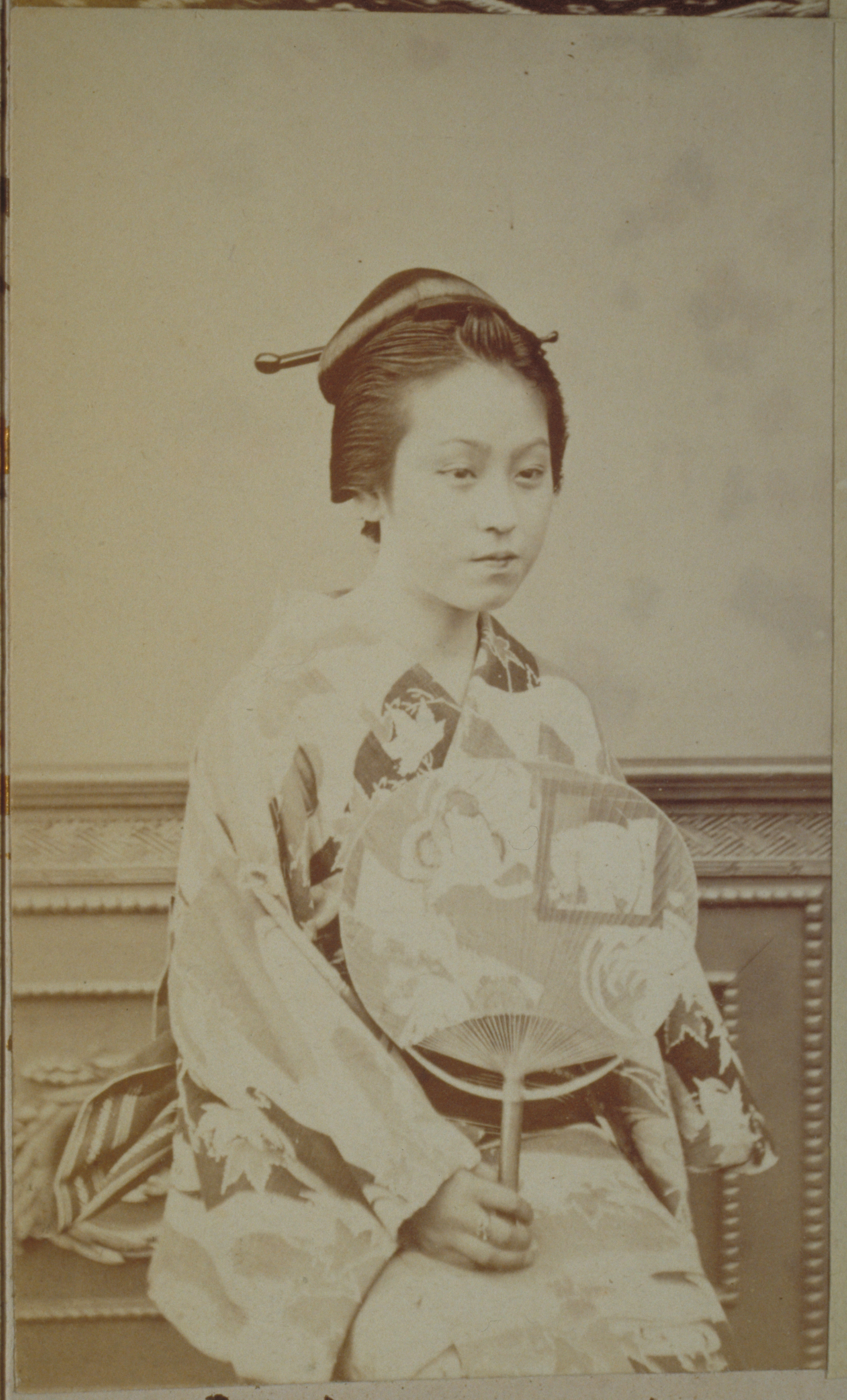 Black-and-white photograph representing a Japanese woman holding a fan and sitting against a wood carved background.