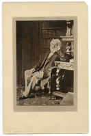 Henry Wadsworth Longfellow seated and leaning his left elbow on Revival style etagere, a dark and heavy piece of furniture. Books on a bookcase form backdrop. Longfellow is wearing formal attire and has ear-length white hair and a full white beard.