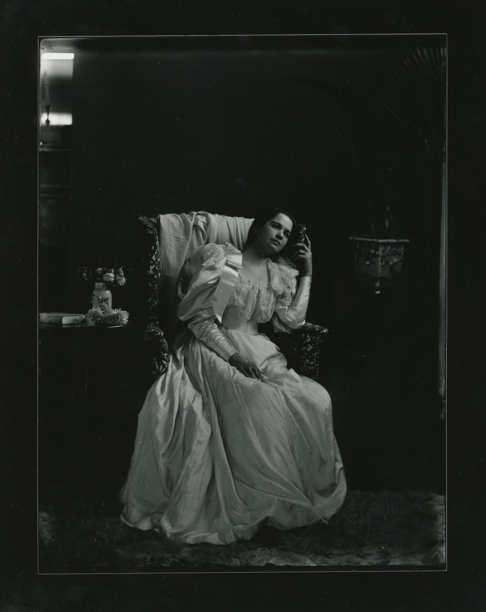 A young white woman dressed in a puffed-sleeve dress sits in an upholstered chair. She rests her head on the left side of the back of the chair.