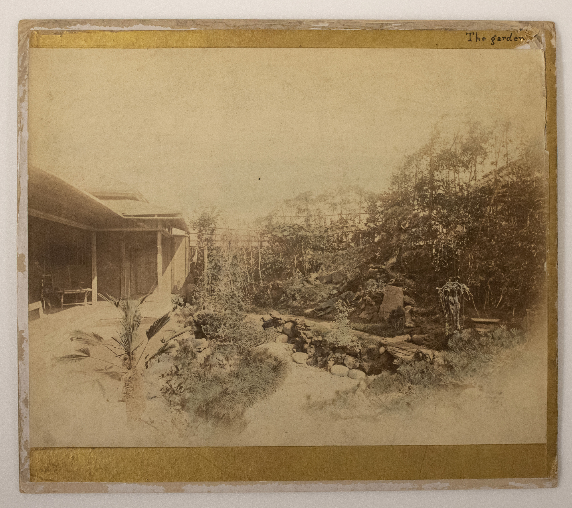 Sepia photograph representing a garden with rocks and plants and a Japanese house on the left. A white man is photographed in the center lying on the grass and reading a book.
