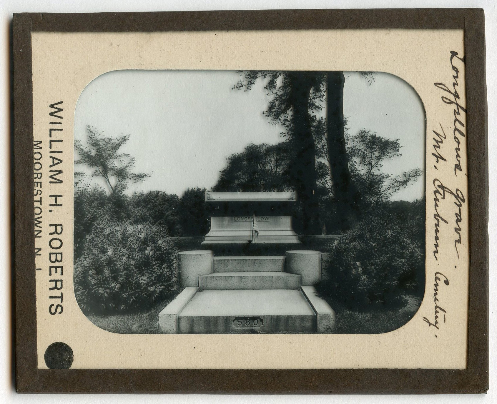 Image of a stone or marble casket-shaped tomb on a raised plot with steps visible in foreground. Bushes flank the plot. Some trees visible in background.