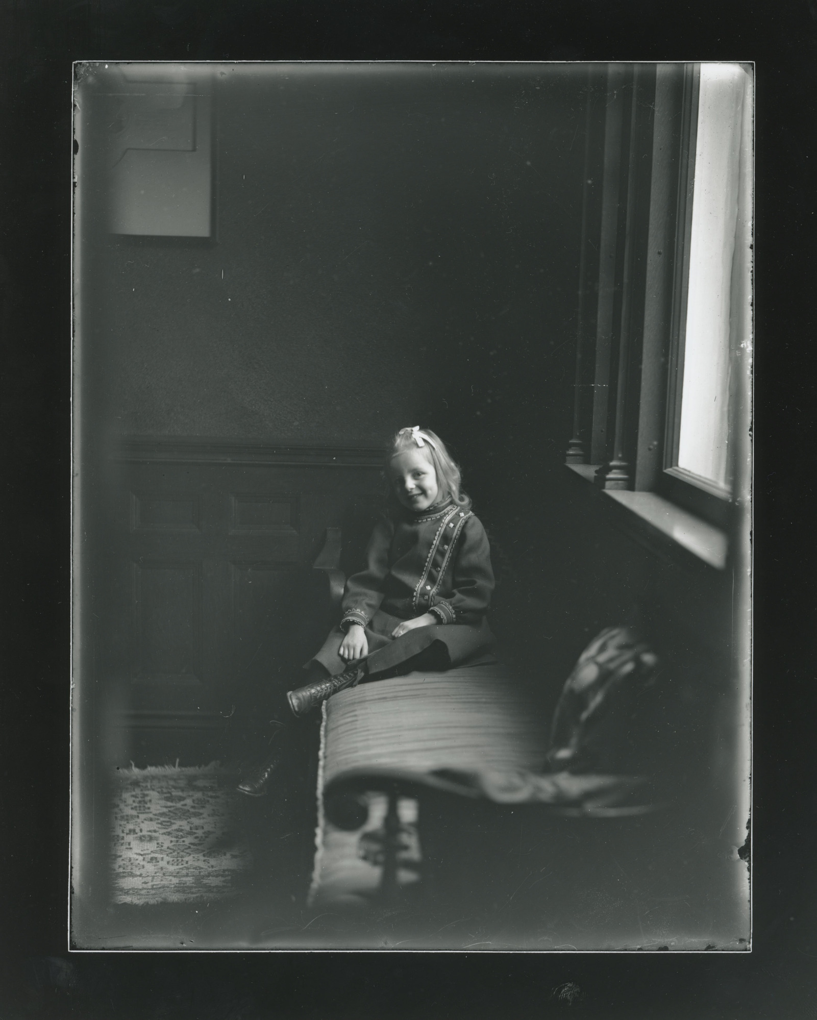 A young white girl sits on a couch underneath a window. She turns towards the camera and rests her left leg on the couch.