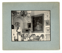 Black and white photograph of the corner of a 19th century parlor, featuring nine paintings and a bust.