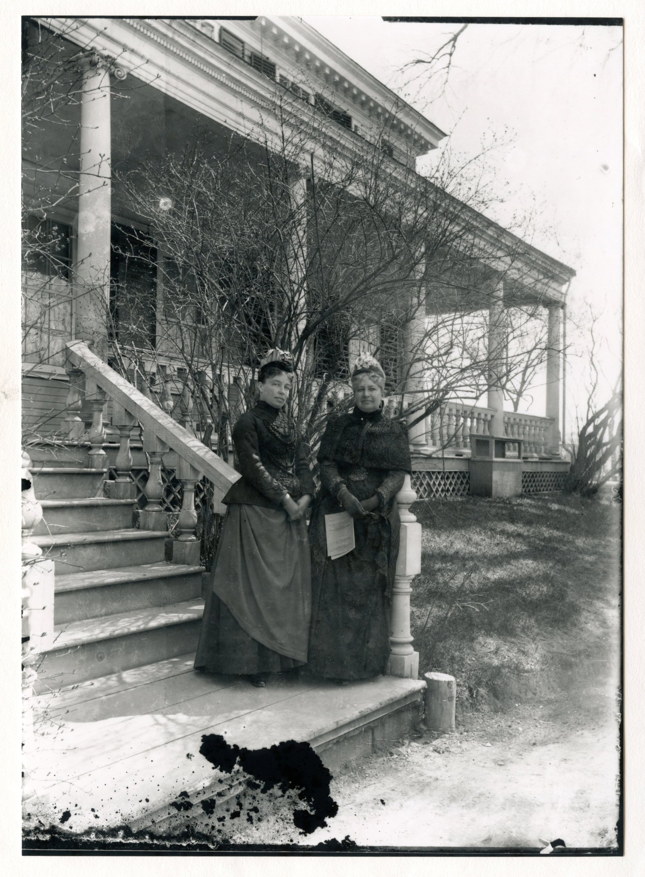 Two white women in dark cold-weather clothing stand at the bottom step of a staircase leading to a porch on the side of a mansion. A bare tree is right behind them.