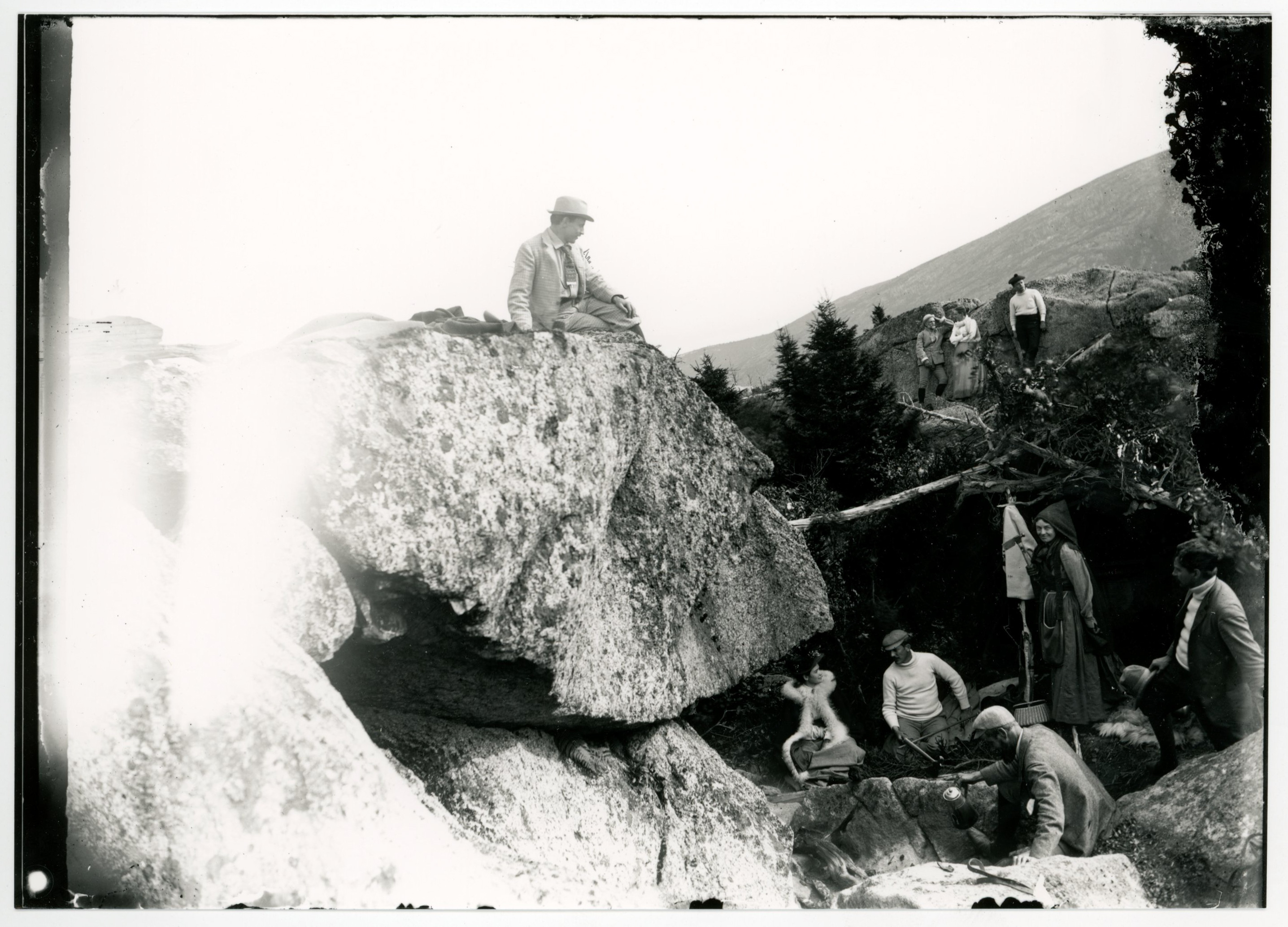 White men and women relax in between and on huge sections of rock. One man carries what seems to be a thermal carafe. A woman stands next to a small wicker basket. One man sits on the highest rock and looks down at the others. Evergreen trees are between the rocks, mountains in the background.