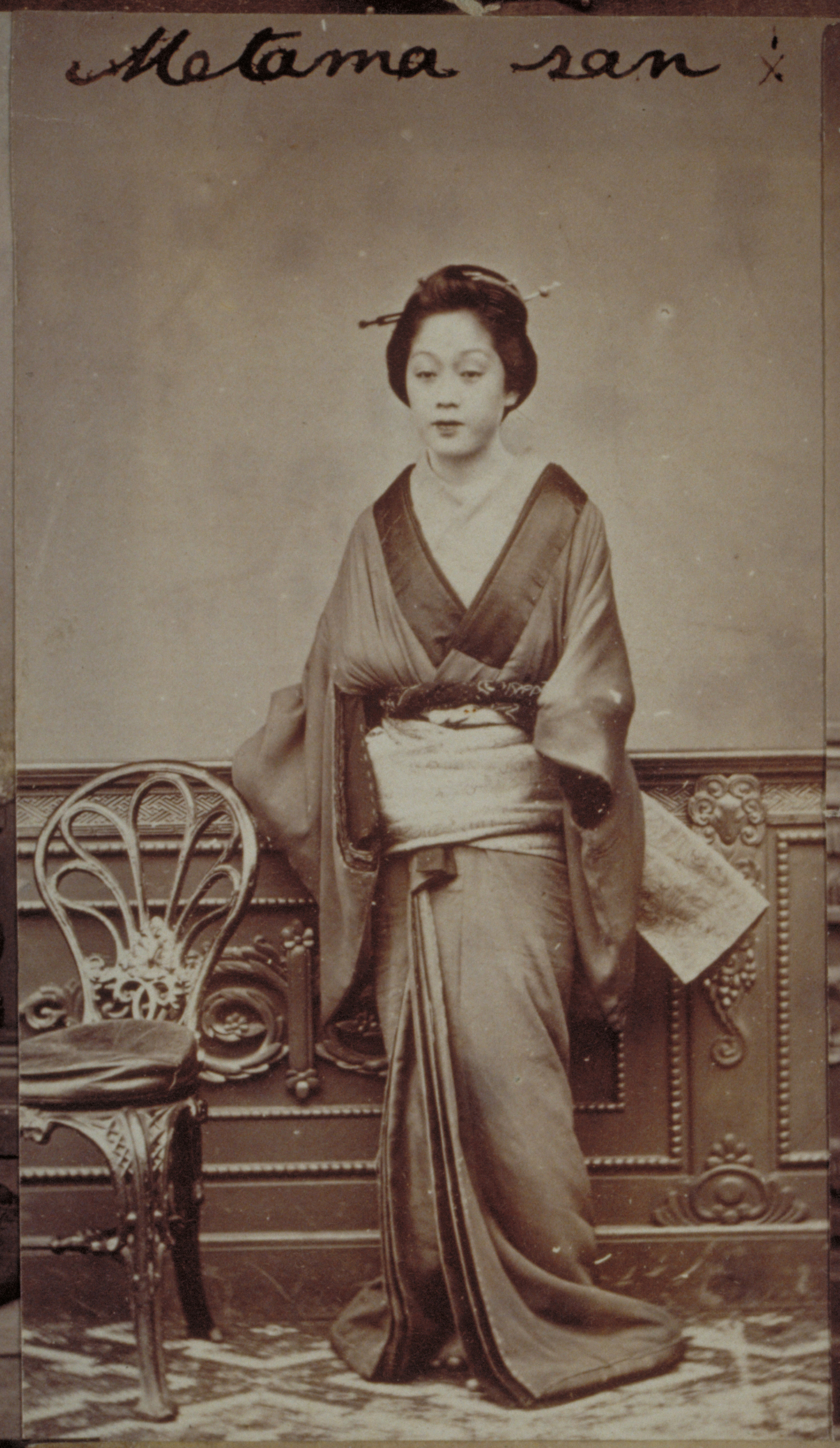 Black-and-white photograph with an inscription representing a Japanese woman standing by a chair against a wood carved wall