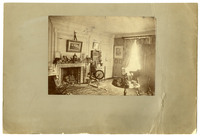 Black and white photograph of Victorian bedroom with fireplace, spinning wheel and bookshelf featured.