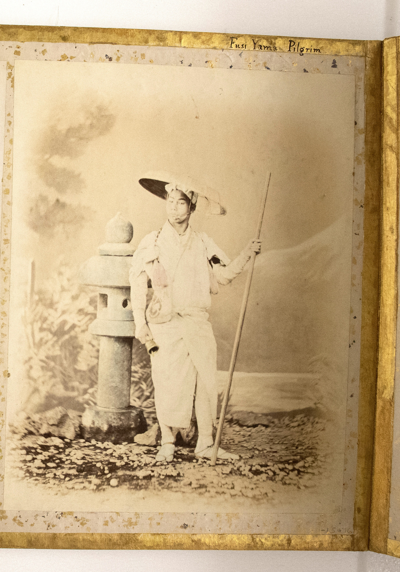 Sepia photograph colored with blue and pink details representing a Japanese man in a hat posing with a staff and a bell in his hands and standing by a stone lantern. The photograph is placed in the center of an album page decorated with a gold-leaf frame, colorful dots, and an inscription.