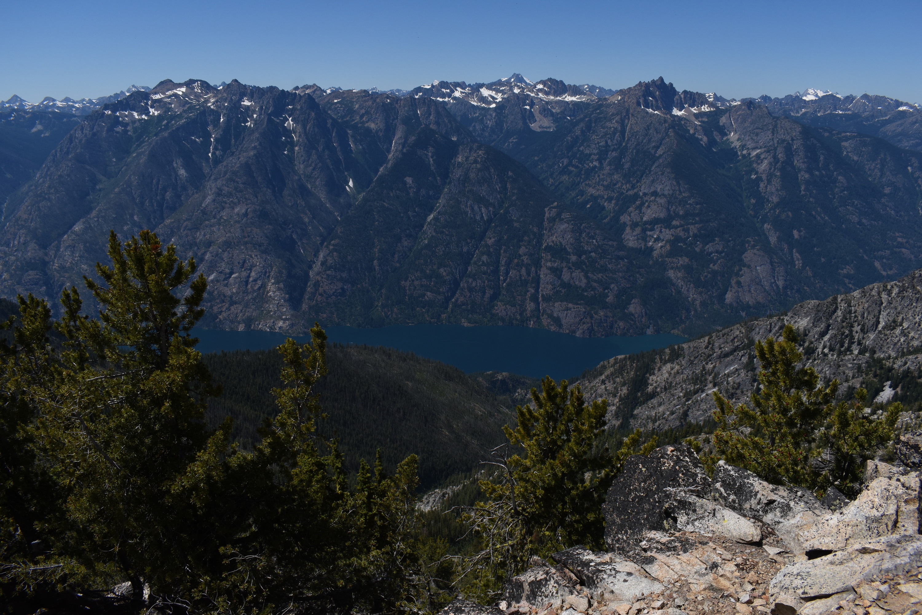 A long thing deep blue lake stretches left to right under steep mountain cliffs as viewed from thousands of feet up.