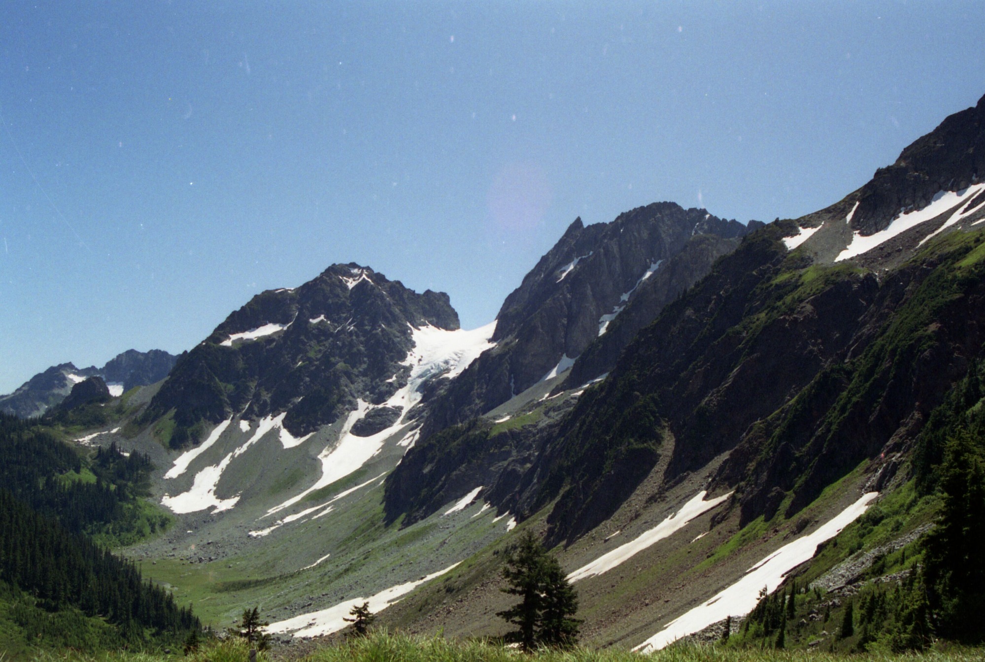 Mountain ridges and peaks covered in snow and grass leading into a grassy basin. On the left side of the image are forested slopes.