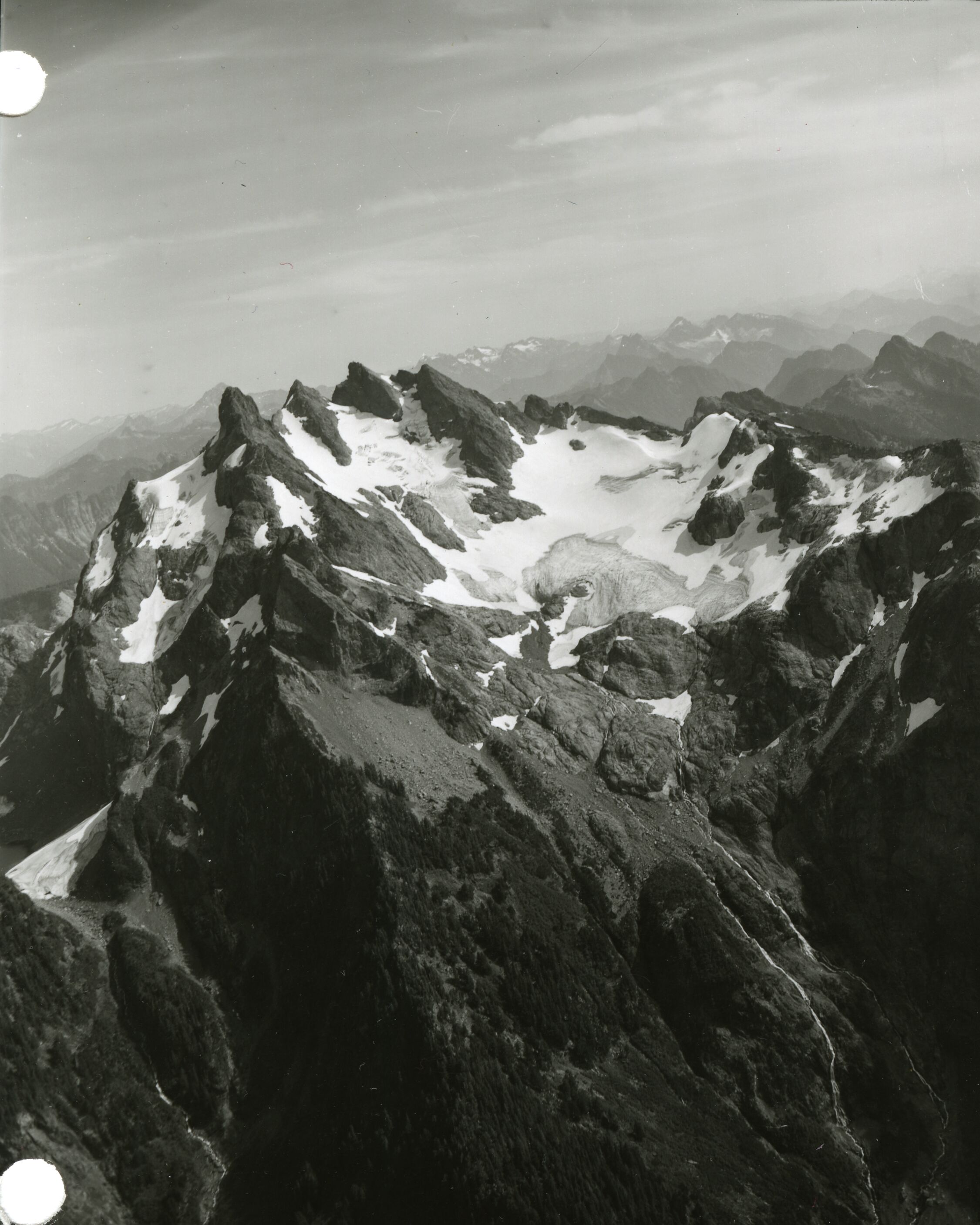 Aerial view of rocky mountans with some snow and ice. More mountains in the background.