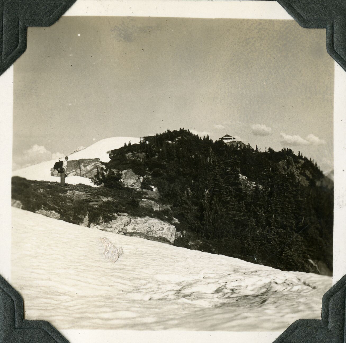 A man with a pack, shown in the distance, standing on a partially snowy ridge, with a snowfield in the foreground.