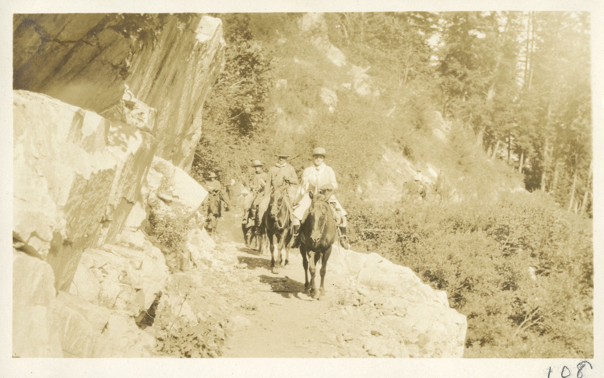 Horseback riders, led by Mary Roberts Rinehart riding, round bend in trail, under sheer rock wall.