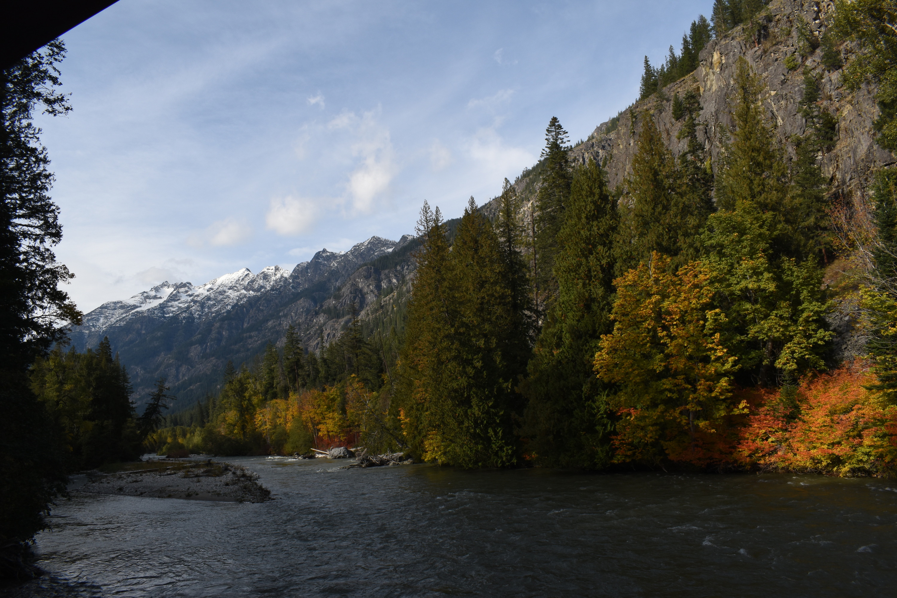 A wide blue river flows through a forest of conifers and maples turning orange with a steep mountain in the background.