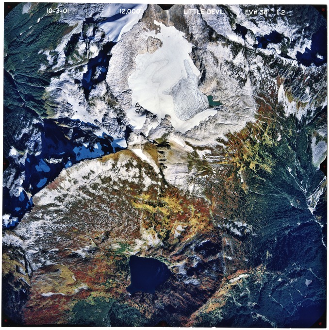 Color aerial photograph of glaciated mountain peak with alpine lake, forested valleys below.