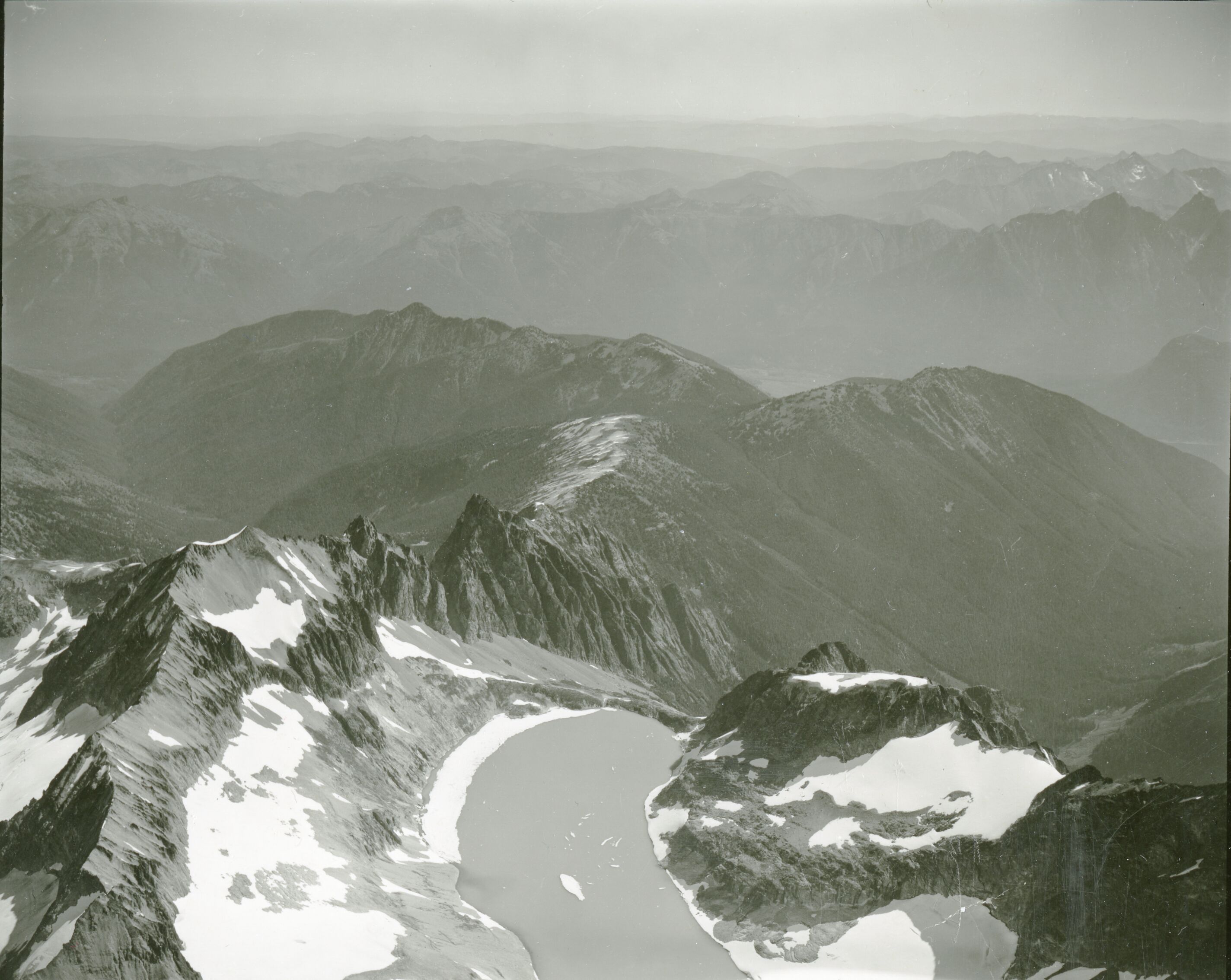 Rocky peaks stretching into the horizon. The closest to the foreground has snow and ice clinging to the sides and has a small lake of melt water at the bottom.