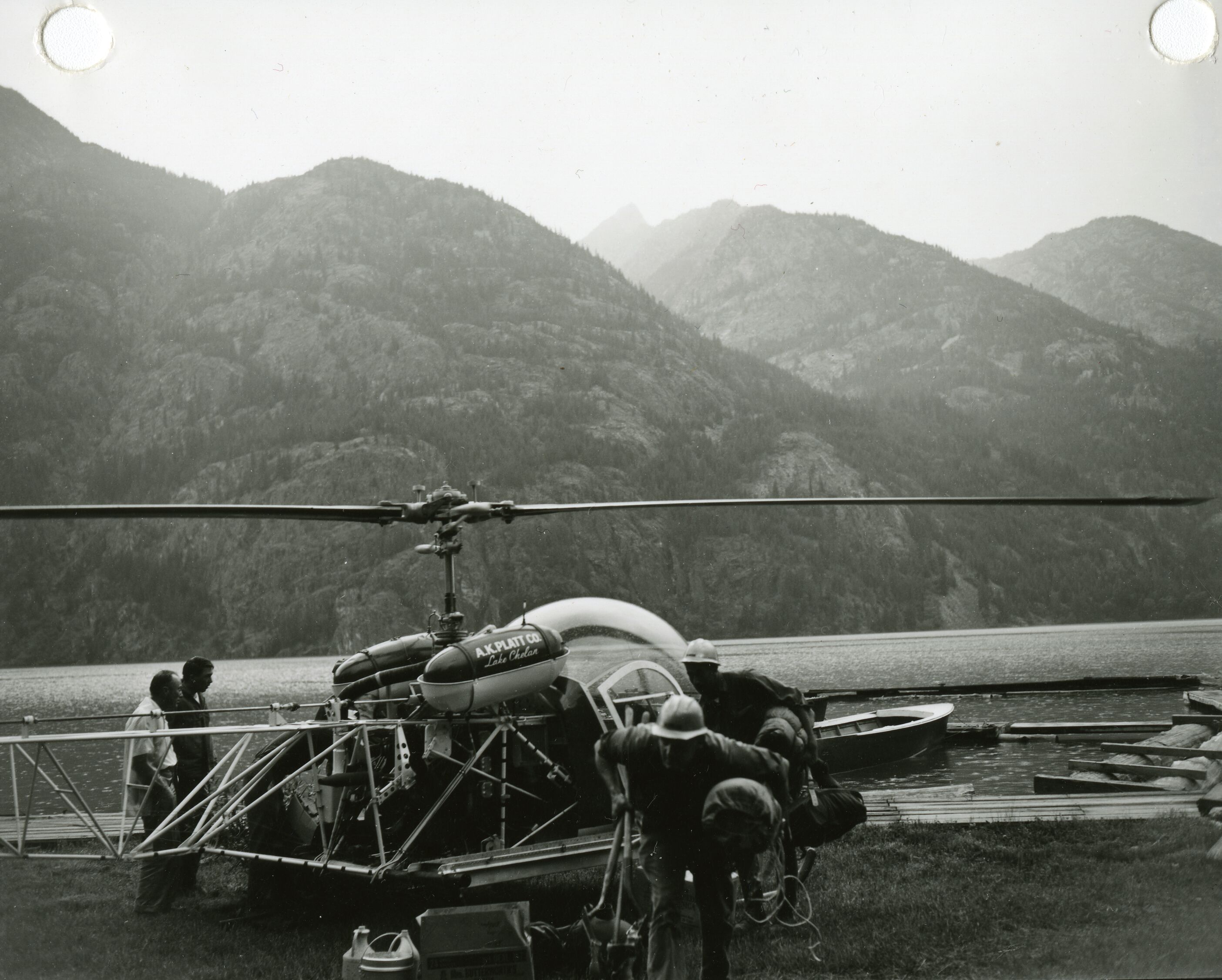 Two people leaving a helicopter with a lake and mountain ridges in the background.