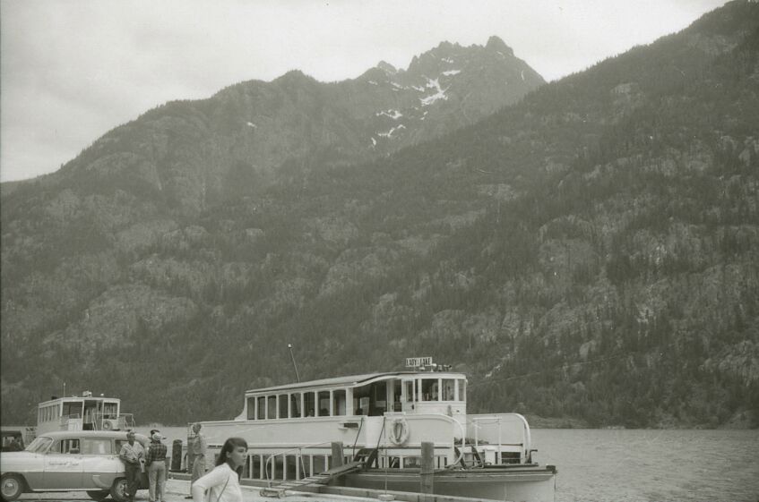 A boat on a lake, cars, and people in front of a wooded slope.