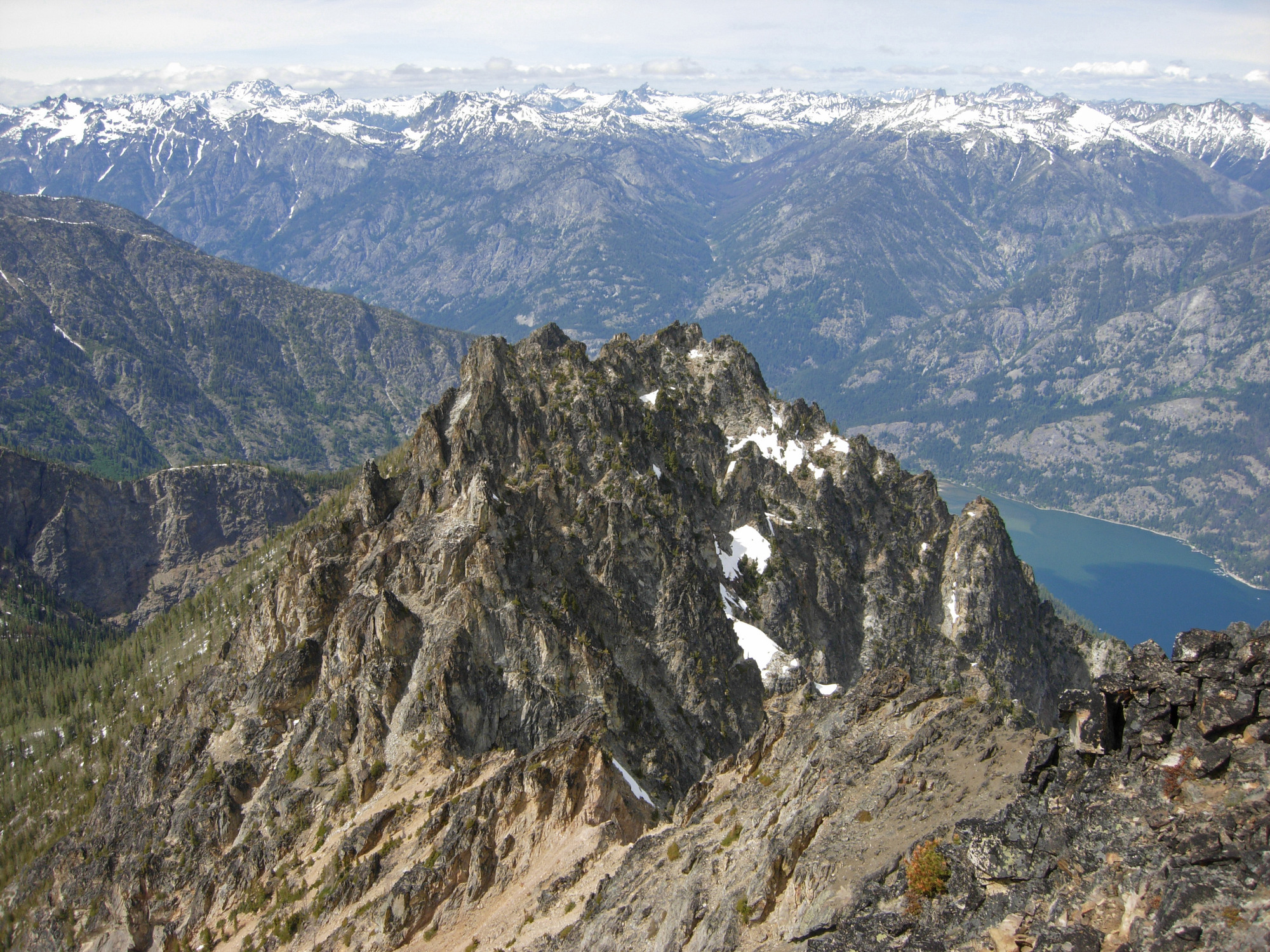 A nearby small craggy peak of brown rock has the backdrop of a range of snow-capped peaks.