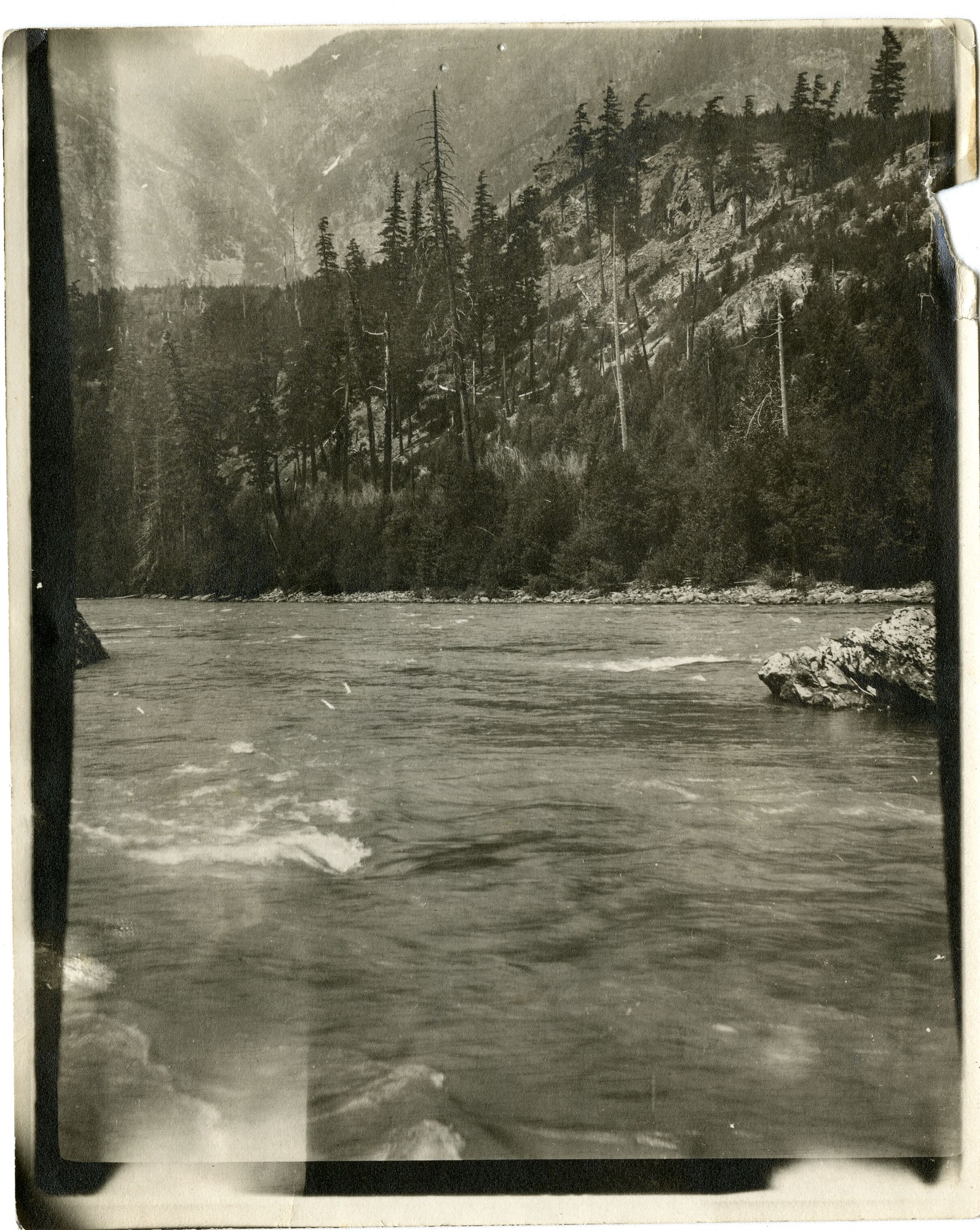 A river is in the foreground and a hill with sparse conifer trees is in the background.