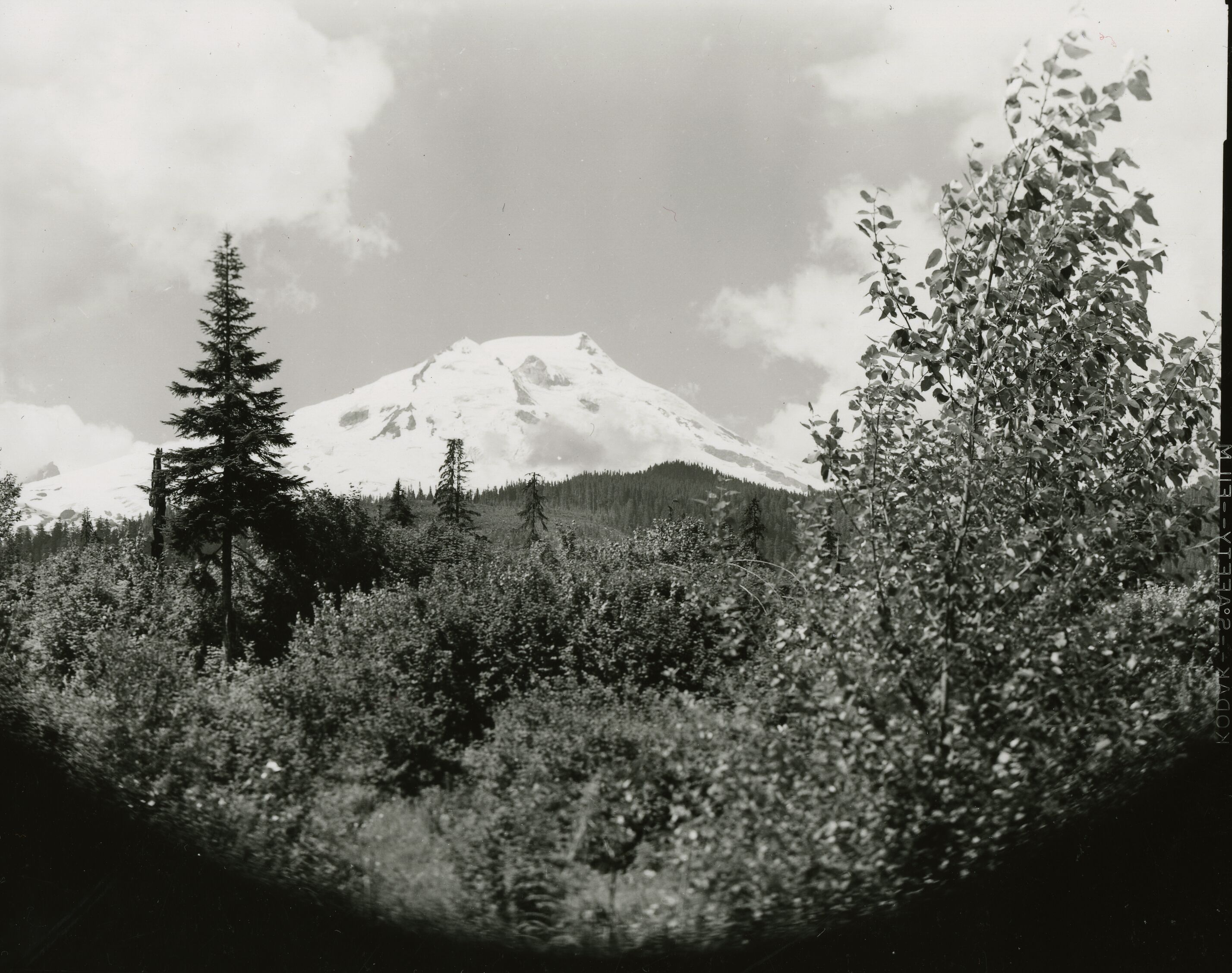 A glaciated peak behind a forested mountain and a lake.