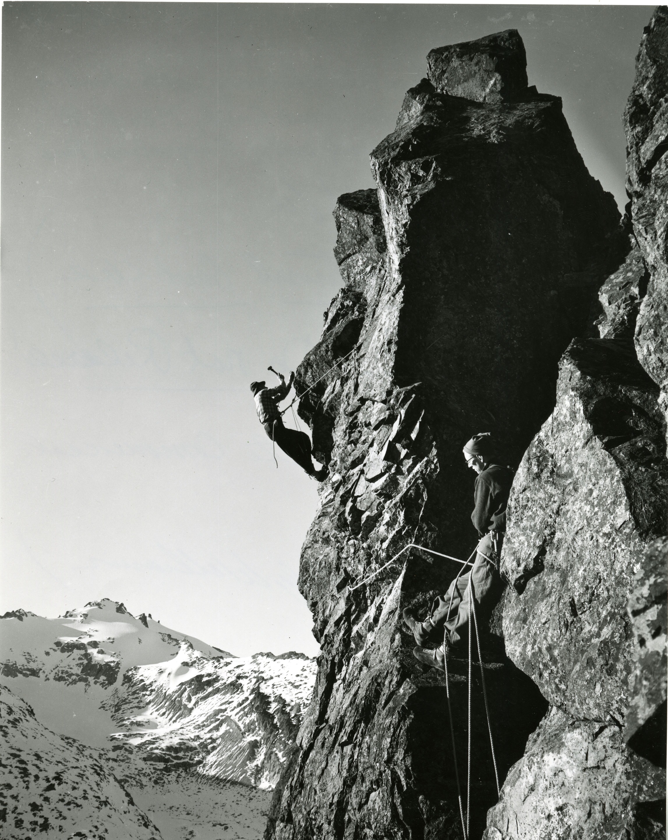 Two men climbing on a steep cliff face with a snow covered mountain peak in the background.