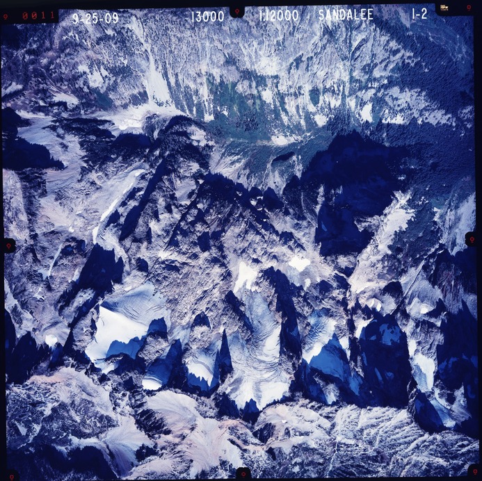 Color aerial photograph of glacier and snowfields on mountain peak with forested valleys below.
