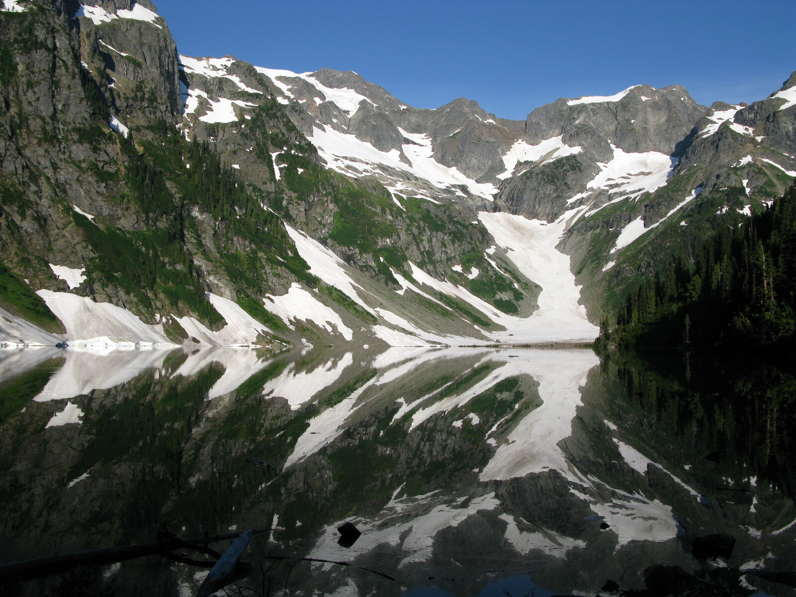 Steep gray cliffs with patches of snow are reflected in still lake water.