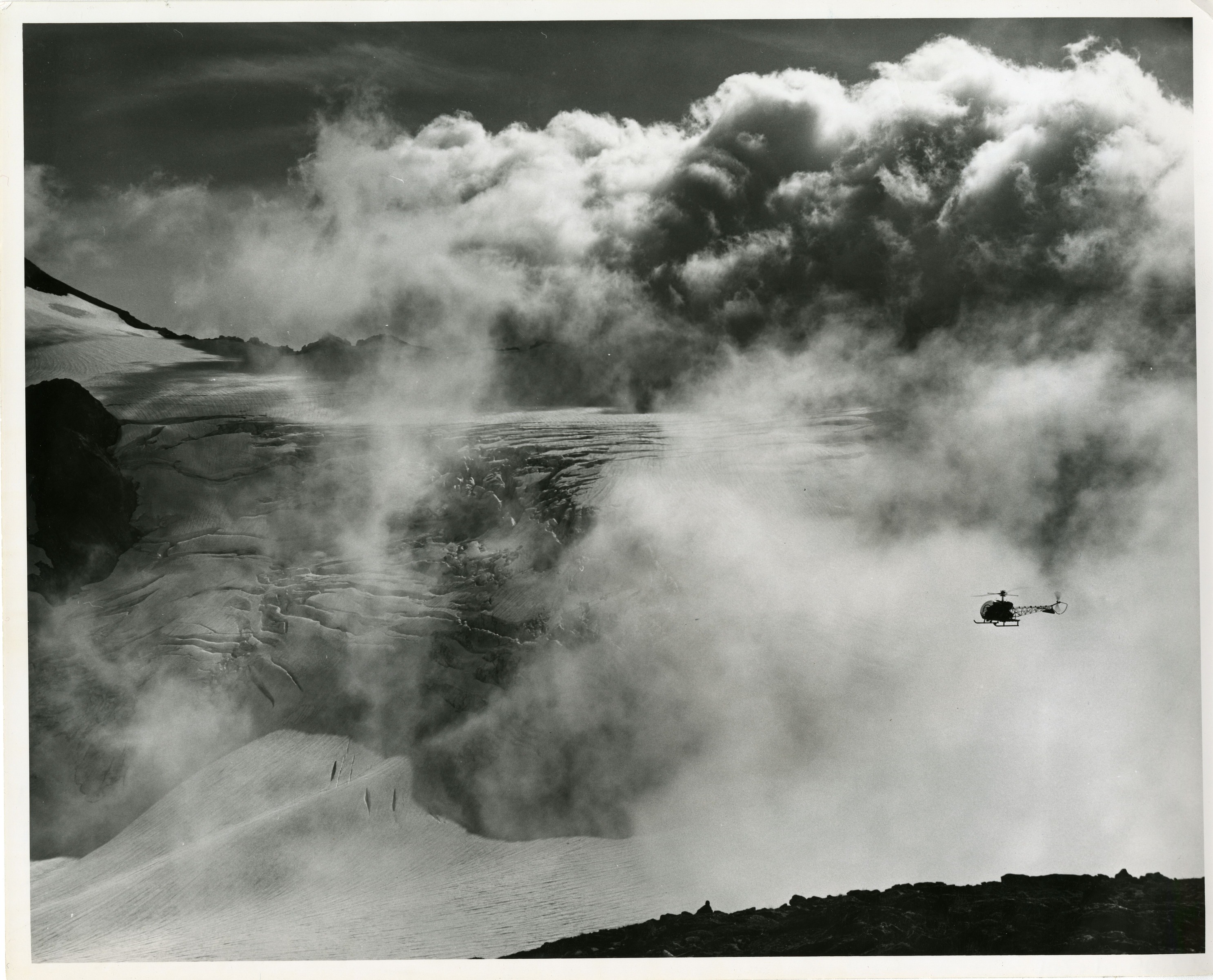 A glacier covered in low clouds, a helicopter in the foreground.