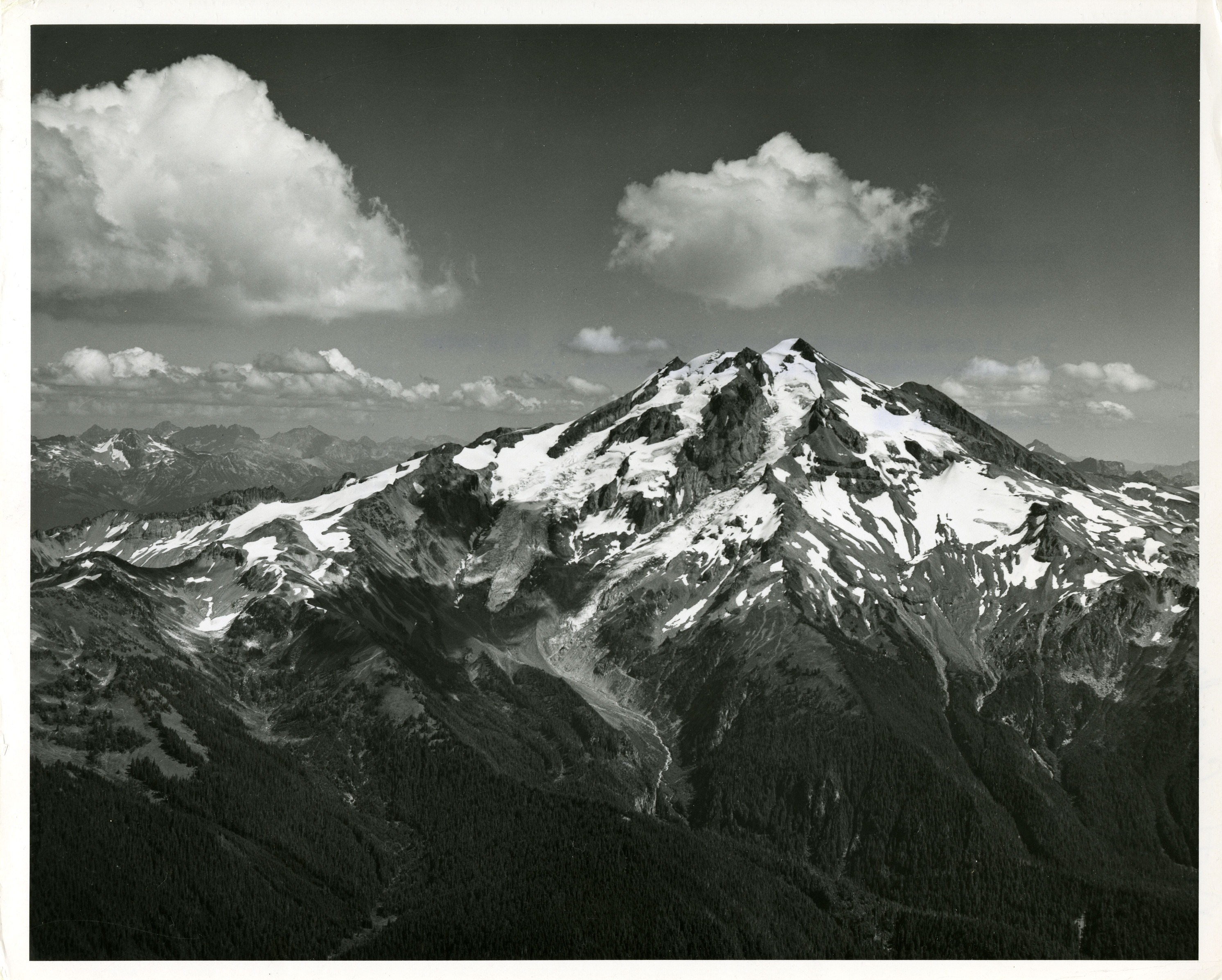 A tall mountain, forested at the bottom. Snow, ice and glaciers start about half-way up. More mountain peaks can be seen in the background.