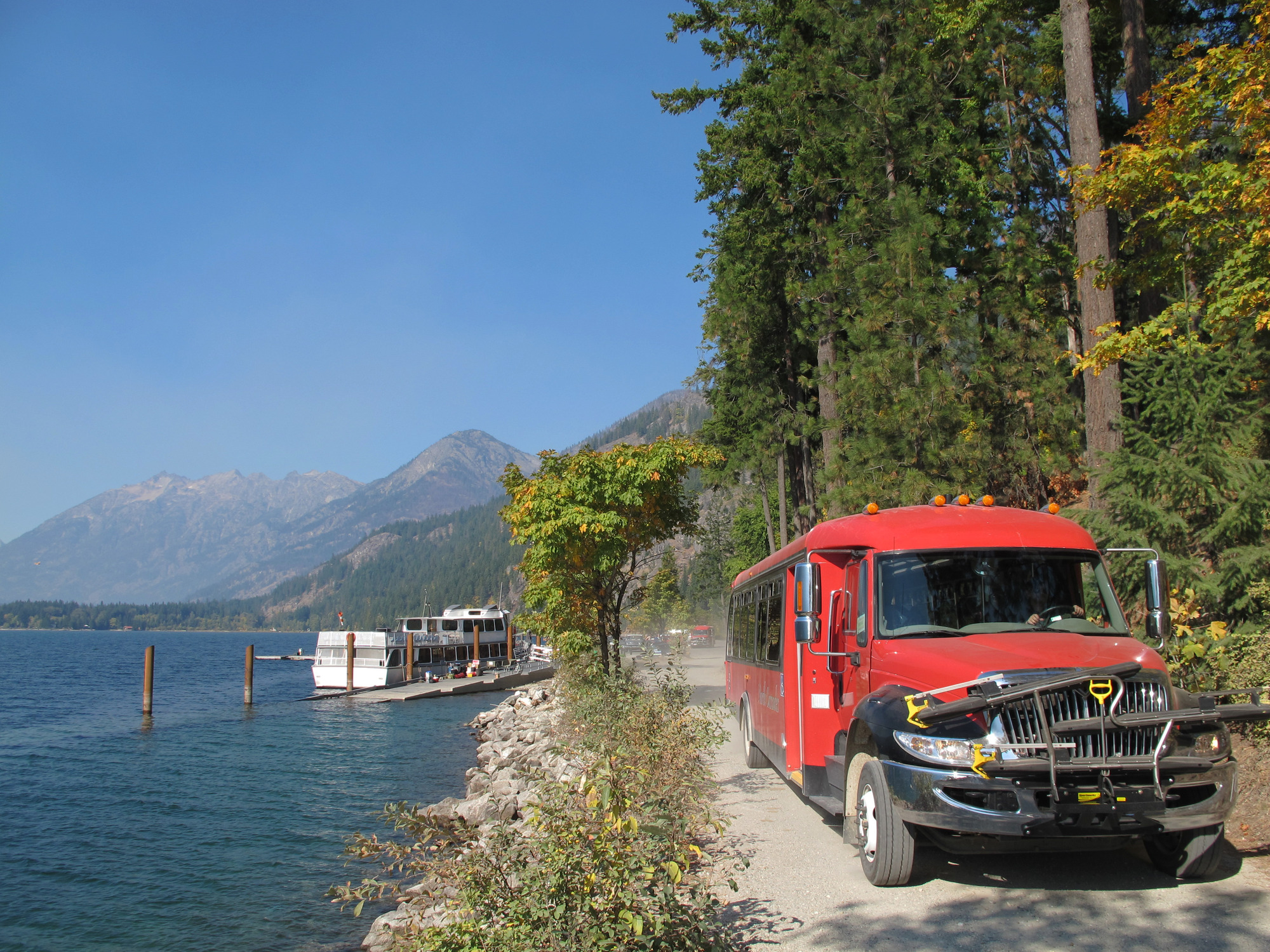A large red bus drives along a forested lakeshore with mountains in the background.