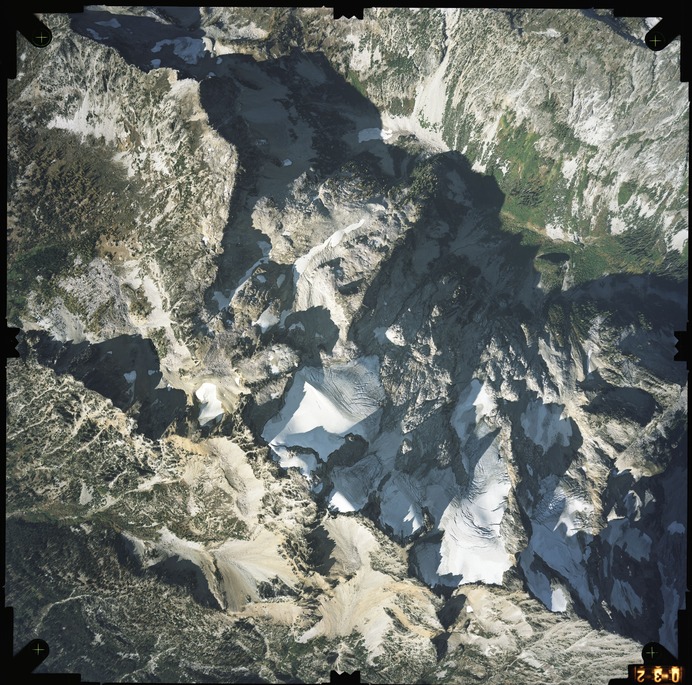 Color aerial photograph of glacier and snowfields on mountain peak with forested valleys below.