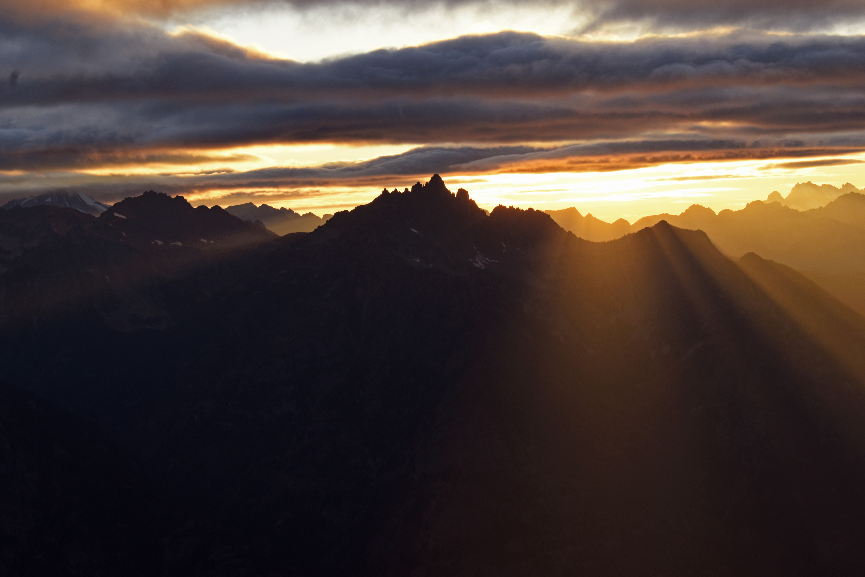 Sun rays shine below gray and orange clouds behind layers of tall mountains.