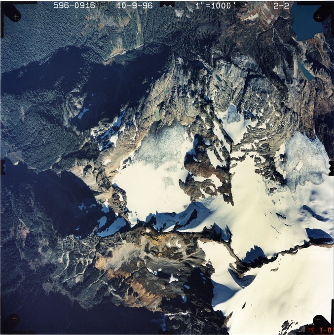 Color aerial photograph of glacier on mountain peak, surrounded by forested valleys.