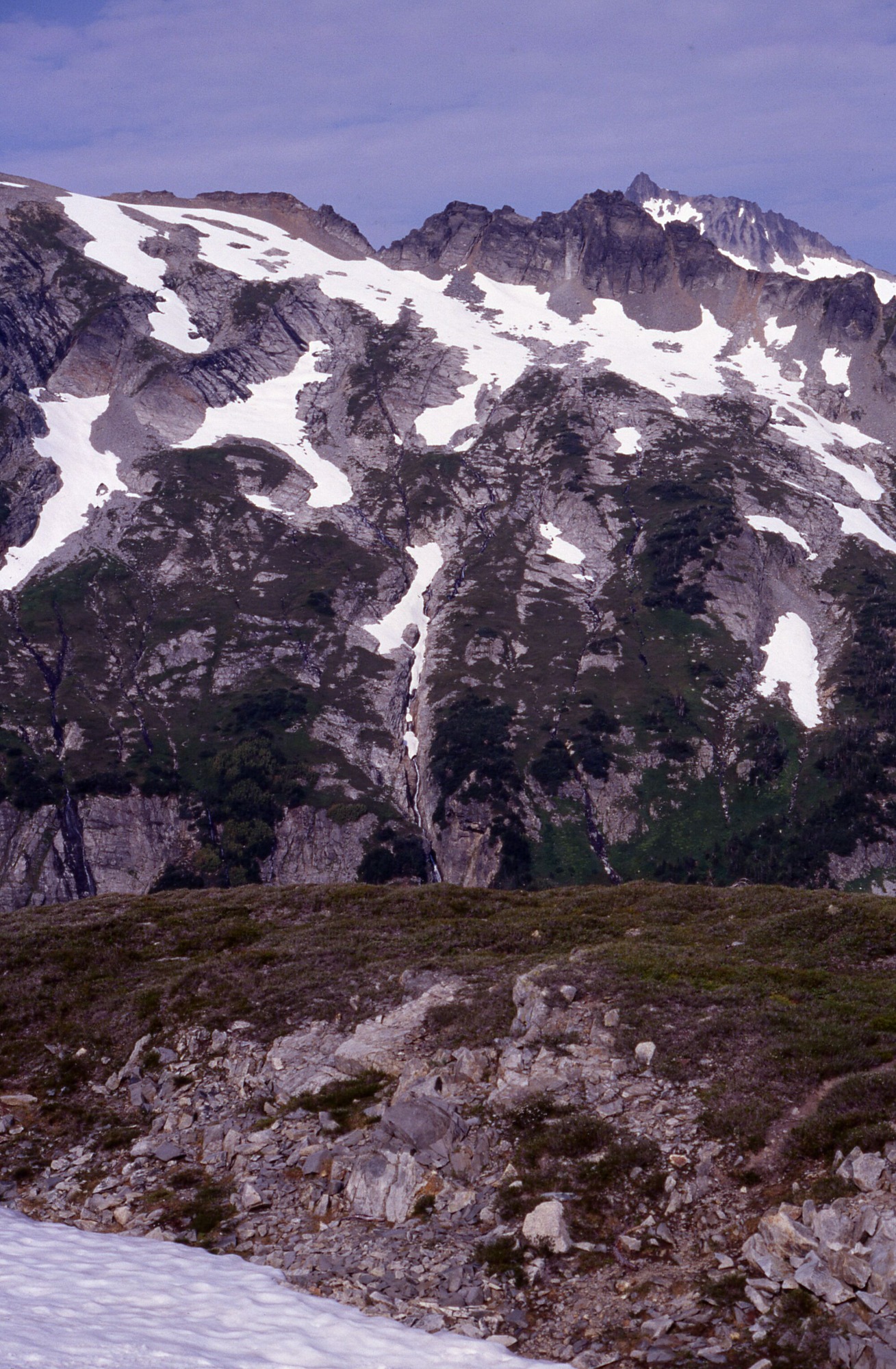 A rocky hill and snow melt. In the distance are snowy mountainsides and peaks.
