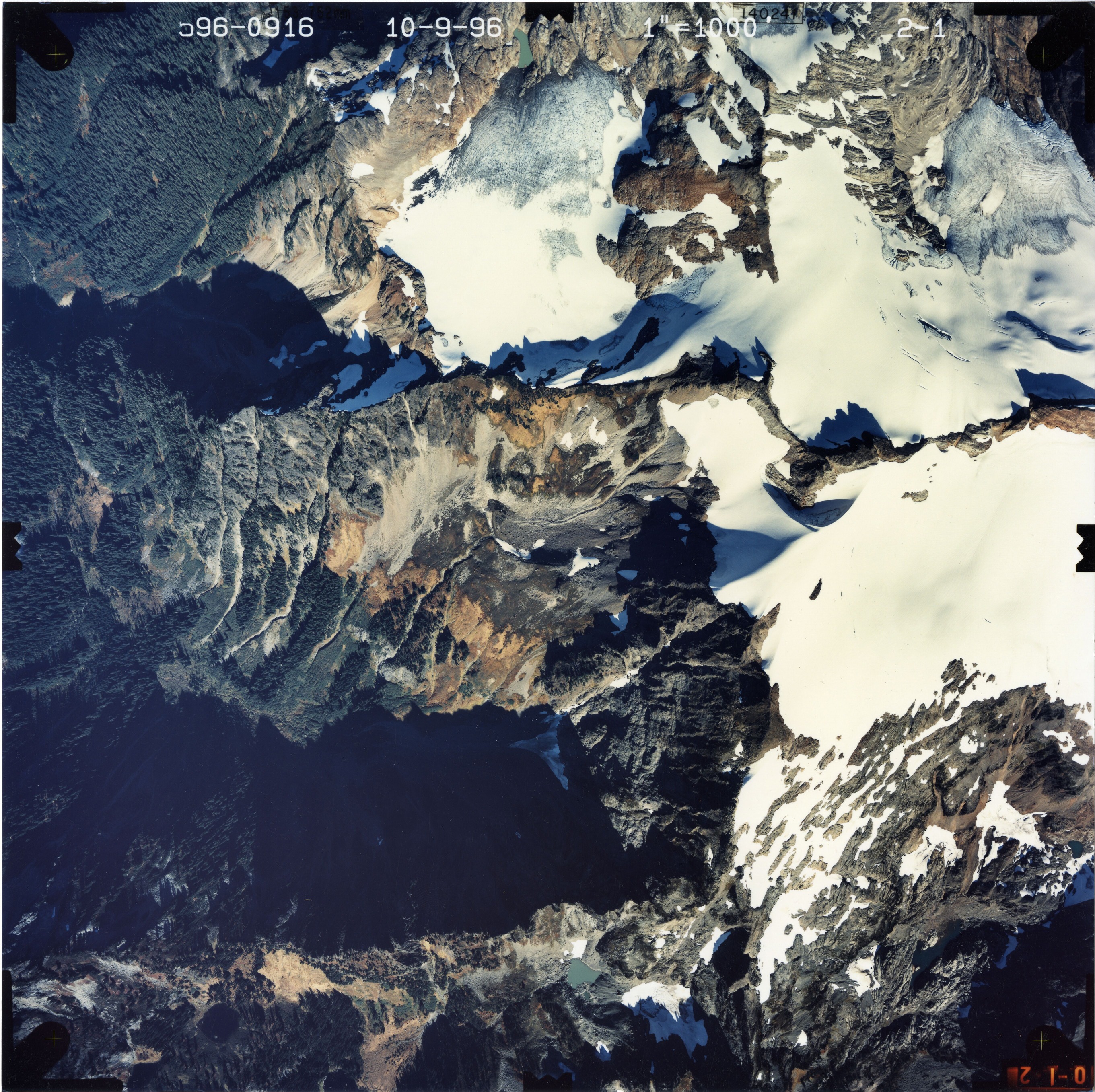 Color aerial photograph of glacier on mountain peak, surrounded by forested valleys.