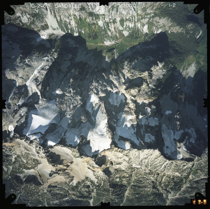 Color aerial photograph of glacier and snowfields on mountain peak with forested valleys below.