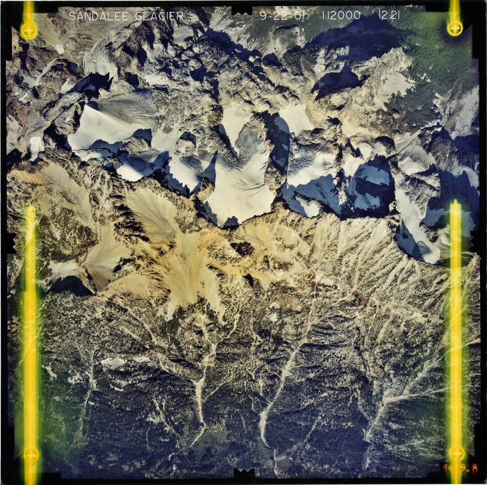 Color aerial photograph of glacier and snowfields on mountain peak with forested valleys below.