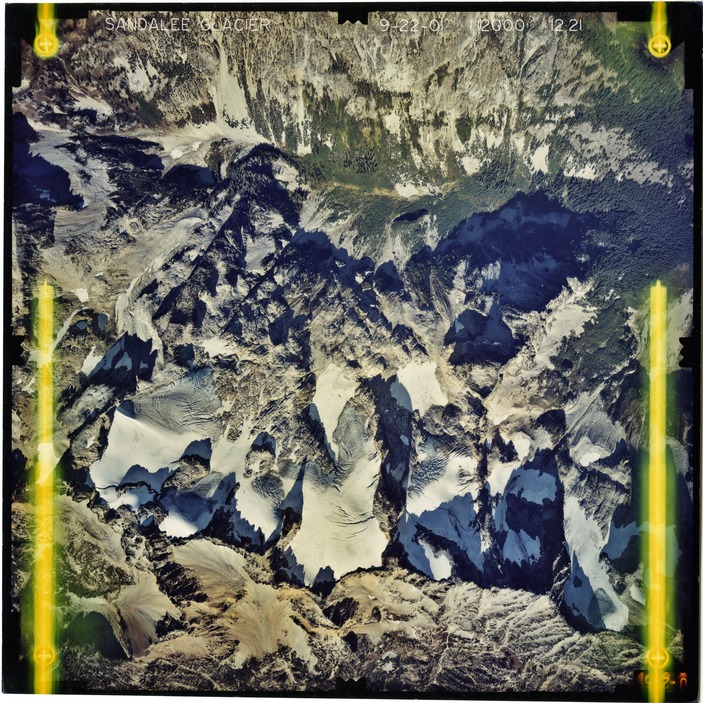 Color aerial photograph of glacier and snowfields on mountain peak with forested valleys below.