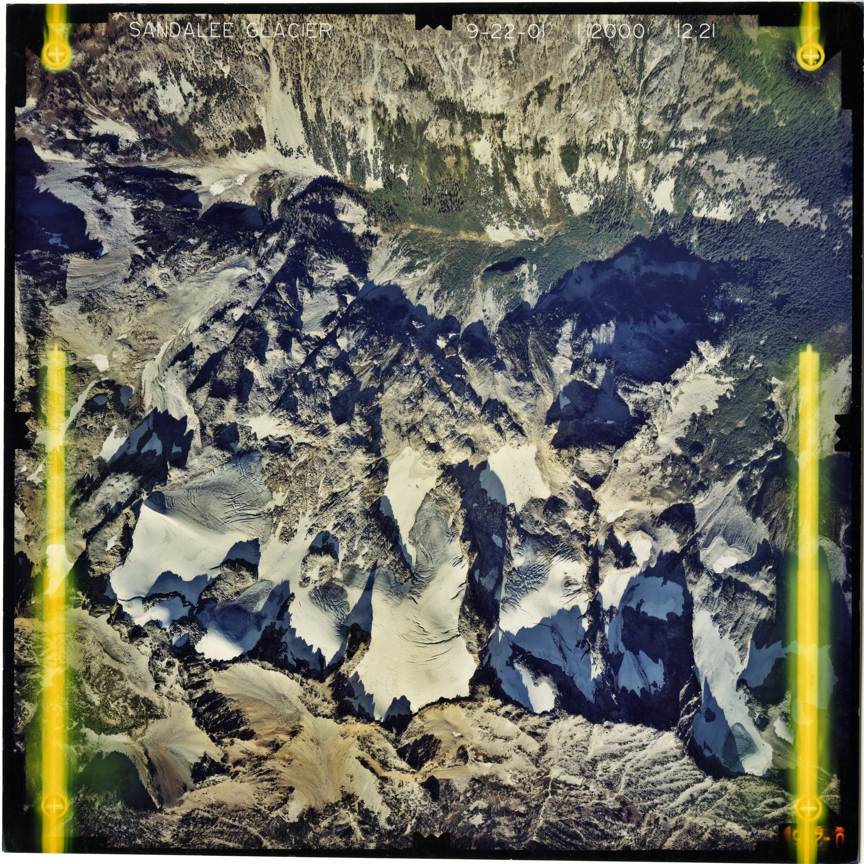Color aerial photograph of glacier and snowfields on mountain peak with forested valleys below.