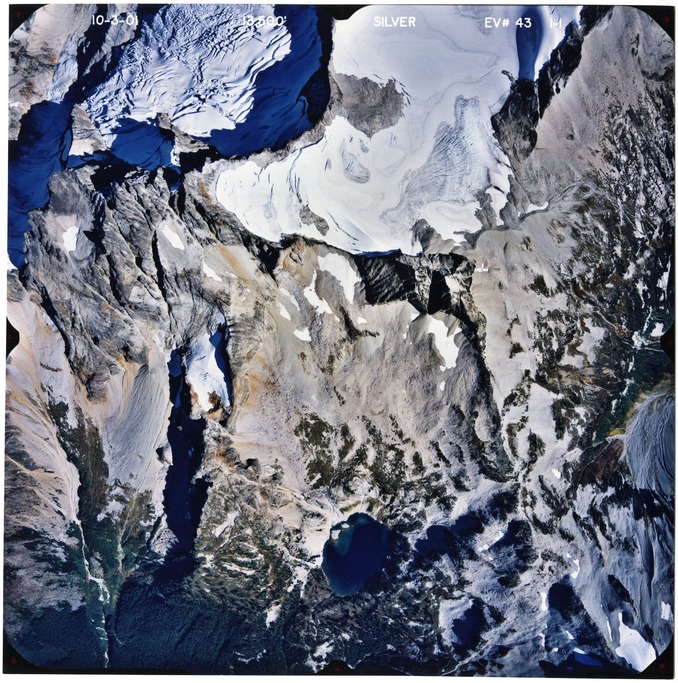Color aerial photograph of glacier, snowfields, and alpine lake on mountain peak with forested valleys below.