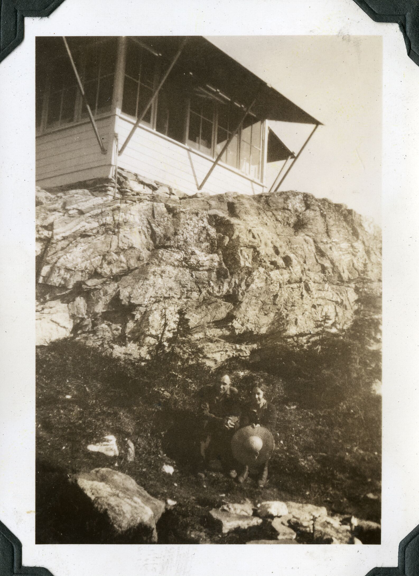 A man and woman sit below a wooden structure atop a rocky outcropping.