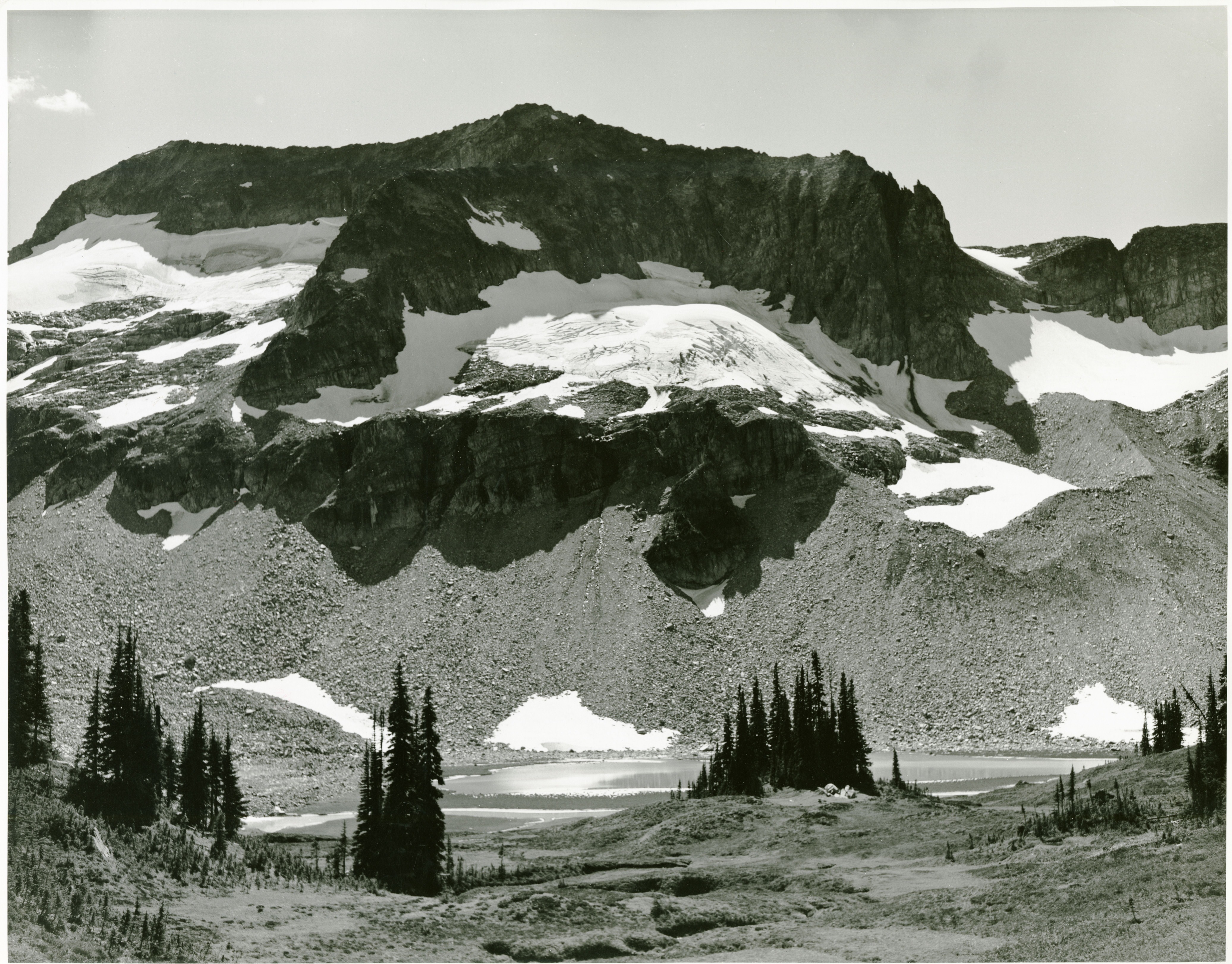 A rocky cliff face above a lake. A meadow is in the foreground, with a handful of coniferous trees.