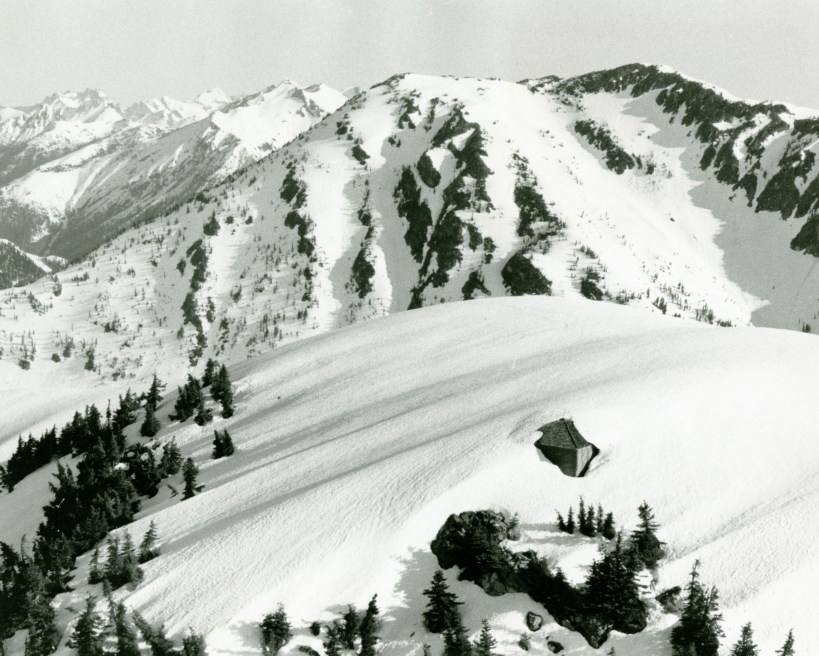 The corner of a small building peaking out from a snow-covered peak.  More snow covered peaks are in the distance.